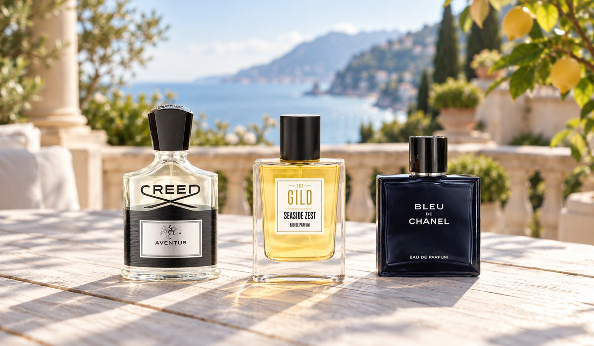 THE GILD Seaside Zest, Creed Aventus and Bleu de Chanel on an outdoor table overlooking a Mediterranean garden and coast.