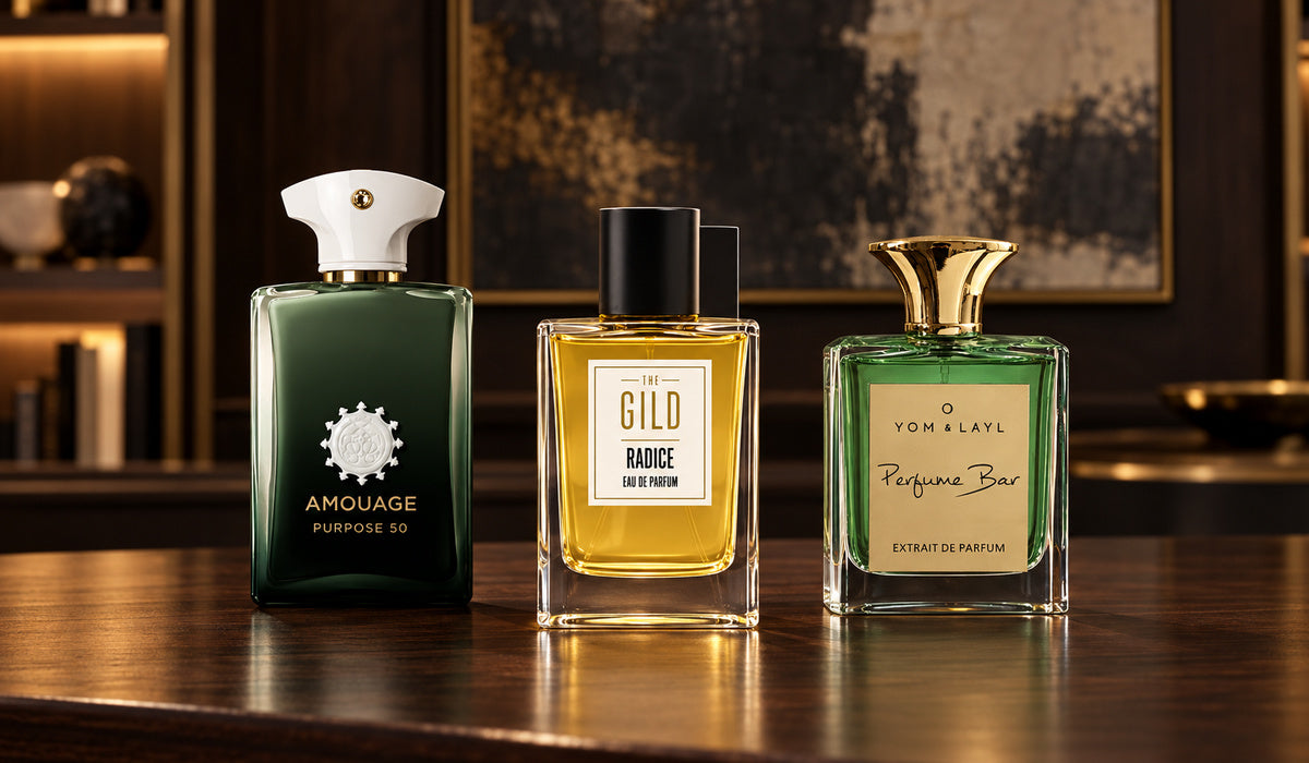 Amouage Purpose 50, THE GILD Radice and Yom & Layl Aim 50 on a dark walnut desk with blurred charcoal, cream and bronze artwork.