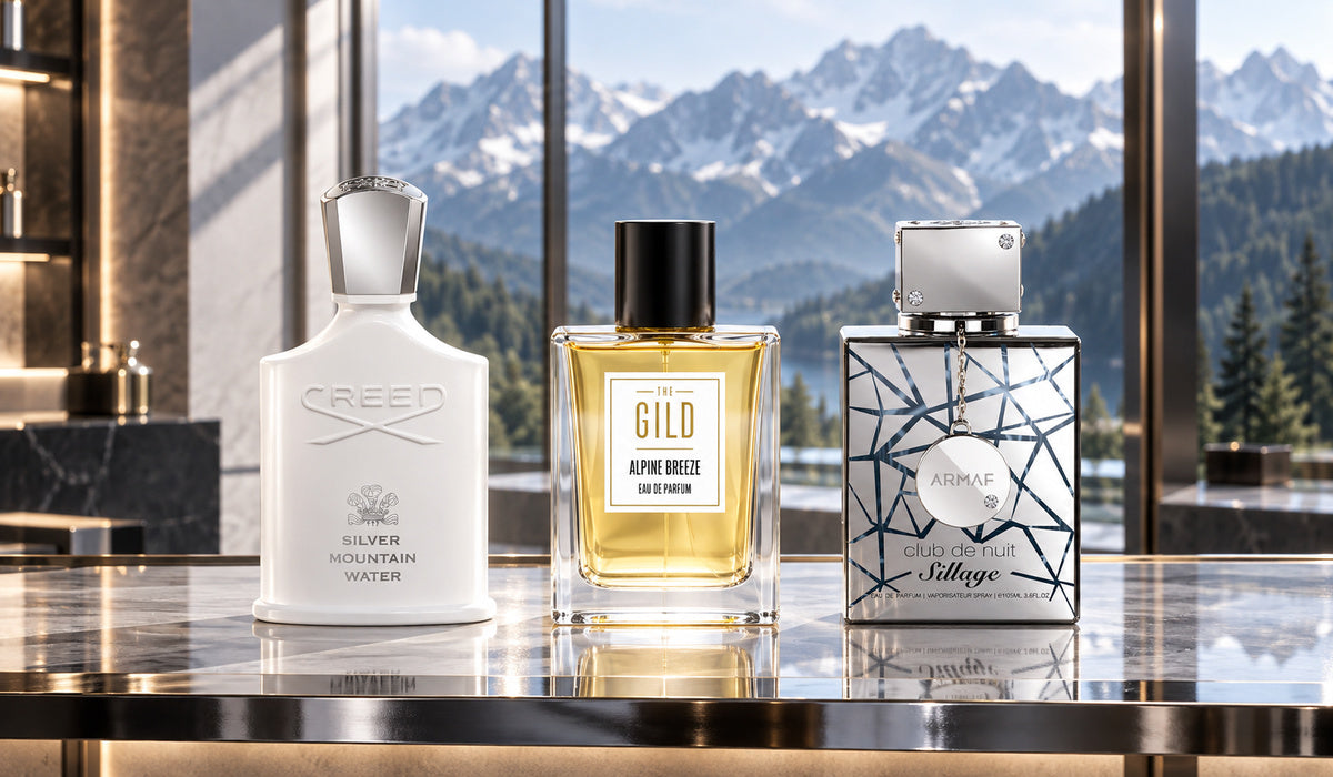 Creed Silver Mountain Water, THE GILD Alpine Breeze and Armaf Club de Nuit Sillage on a silver and glass shelf with softly blurred mountains behind.