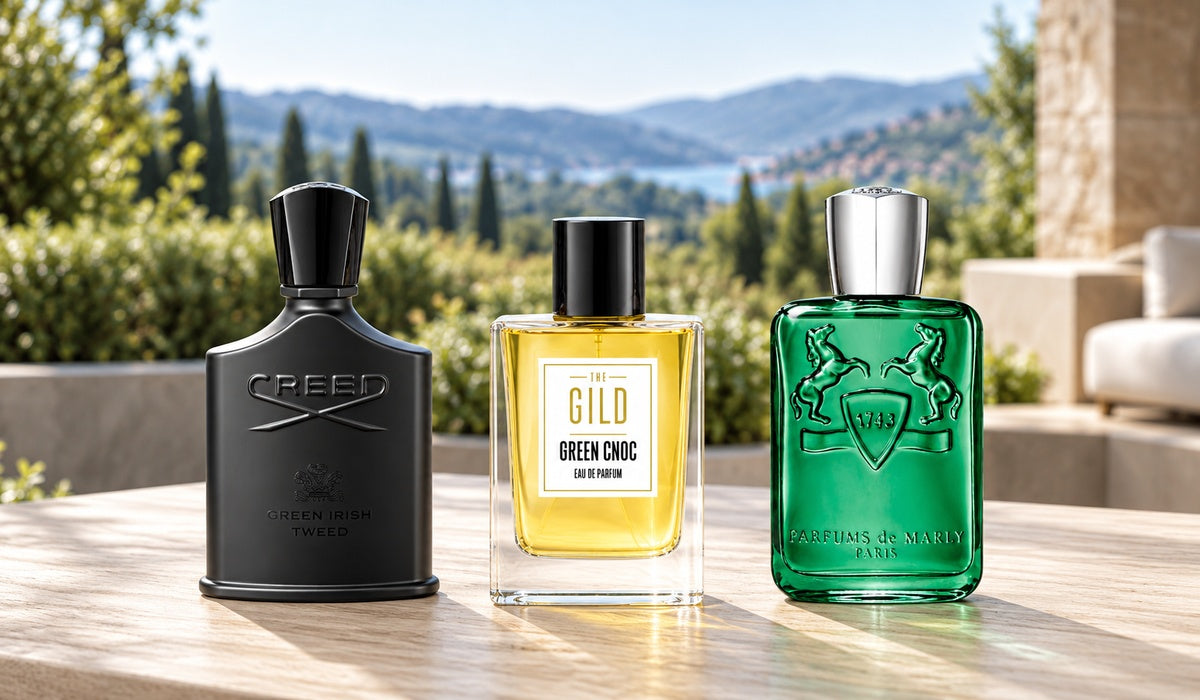 Creed Green Irish Tweed, THE GILD Green Cnoc and Parfums de Marly Greenley on a pale oak terrace table with blurred garden trees and green hills.