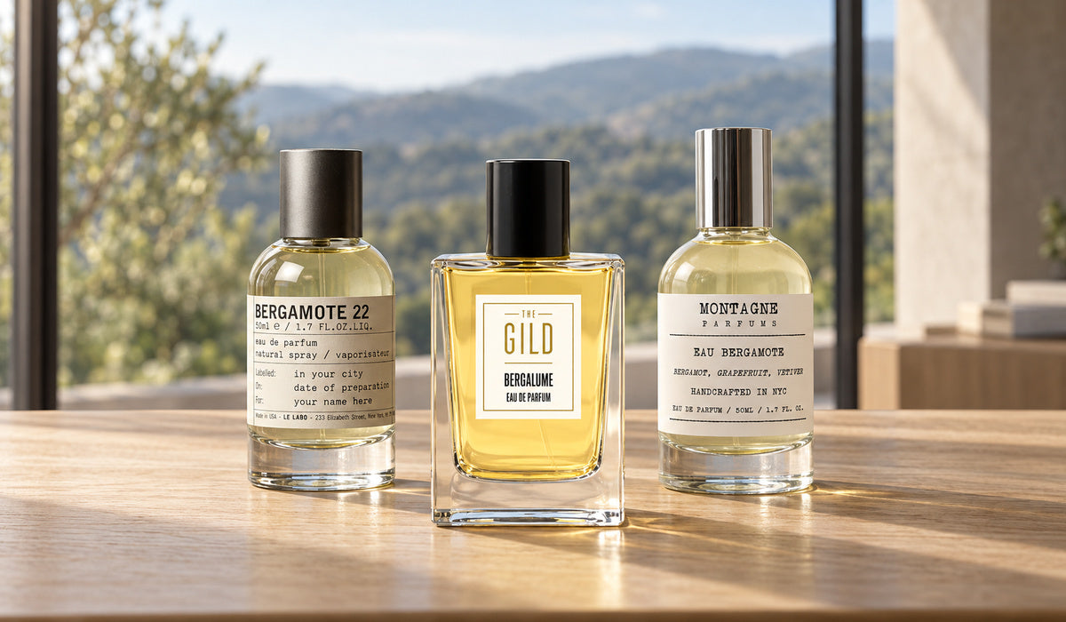 Le Labo Bergamote 22, THE GILD Bergalume and Montagne Eau Bergamote on a light oak table with blurred green hills through a window.