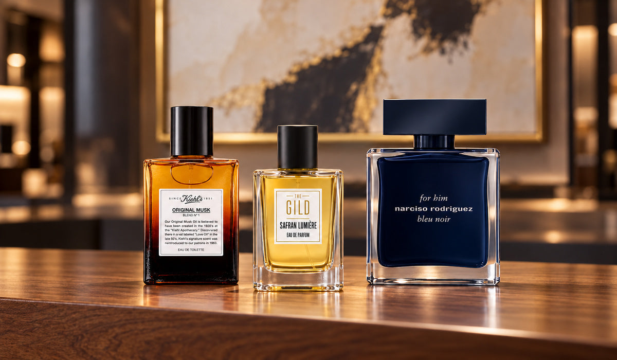 Kiehl's Original Musk, THE GILD Safran Lumière and Narciso Rodriguez for him bleu noir on a walnut retail counter with a blurred cream painting in a gold frame.