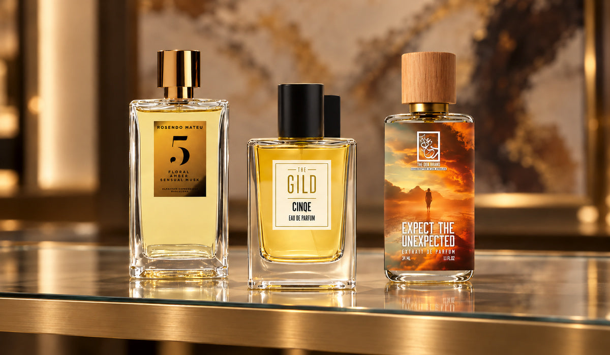Rosendo Mateu No. 5, THE GILD Cinqe and DUA Expect the Unexpected on a glass retail shelf with blurred brown, cream and gold artwork.
