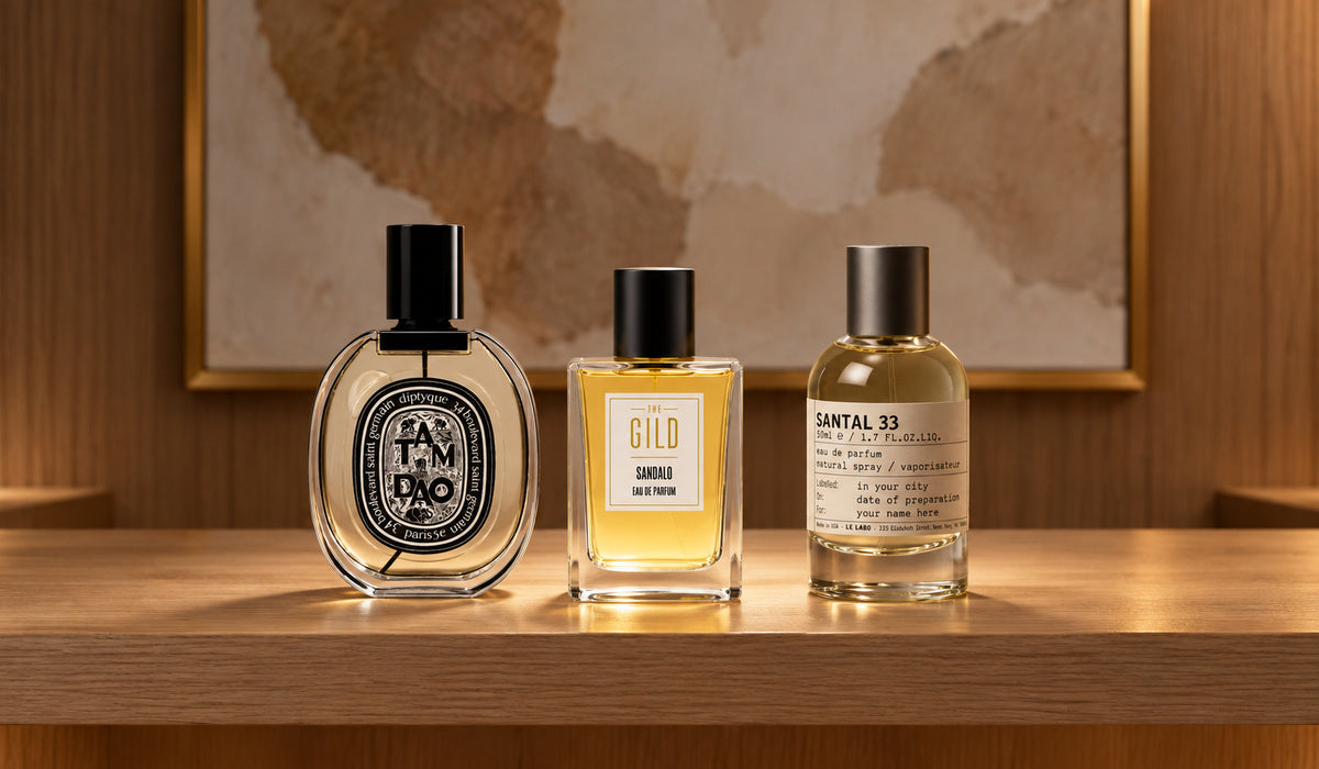 Diptyque Tam Dao, THE GILD Sandalo and Le Labo Santal 33 on a light oak retail shelf with blurred cream and tan artwork.