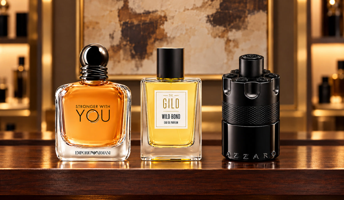 Emporio Armani Stronger With You, THE GILD Wild Bond and Azzaro The Most Wanted Eau de Parfum Intense on a mahogany shelf with blurred cream and brown artwork.