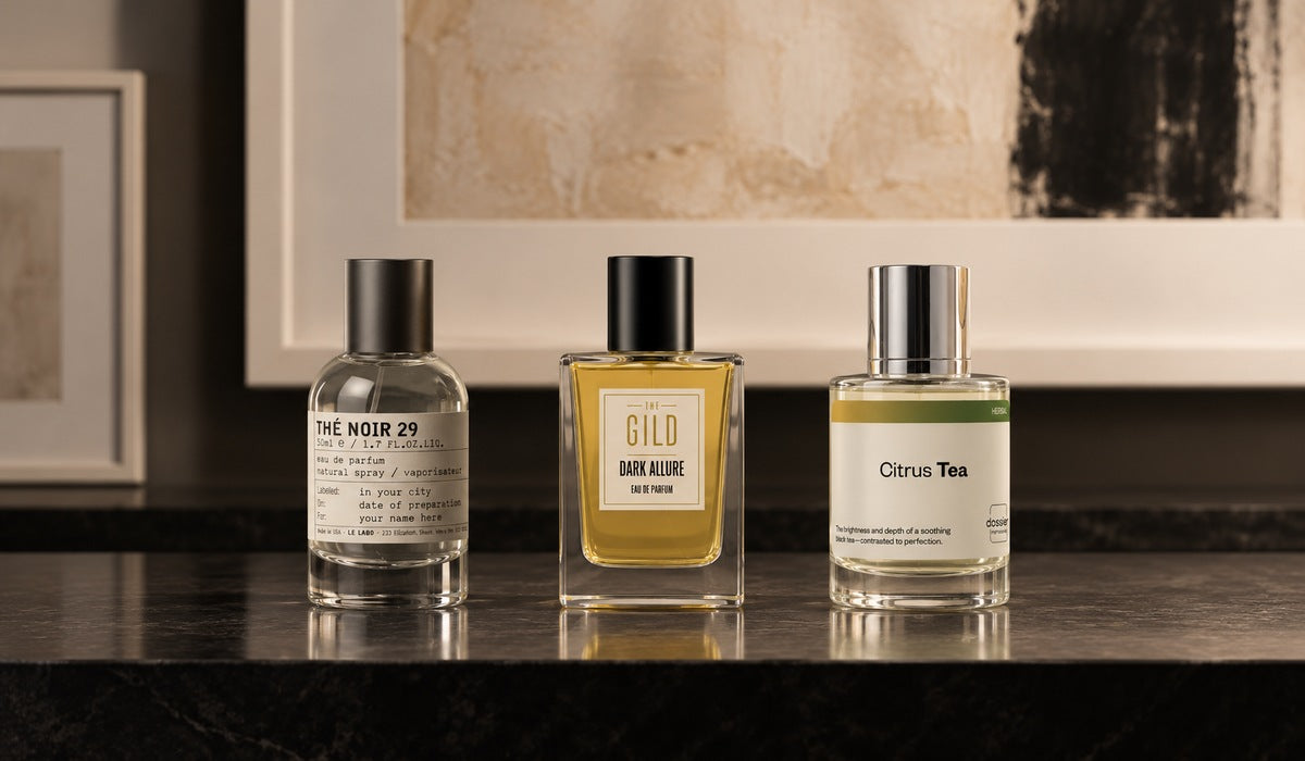 Le Labo Thé Noir 29, THE GILD Dark Allure and Dossier Citrus Tea on a black shelf with white frames and a blurred cream painting.